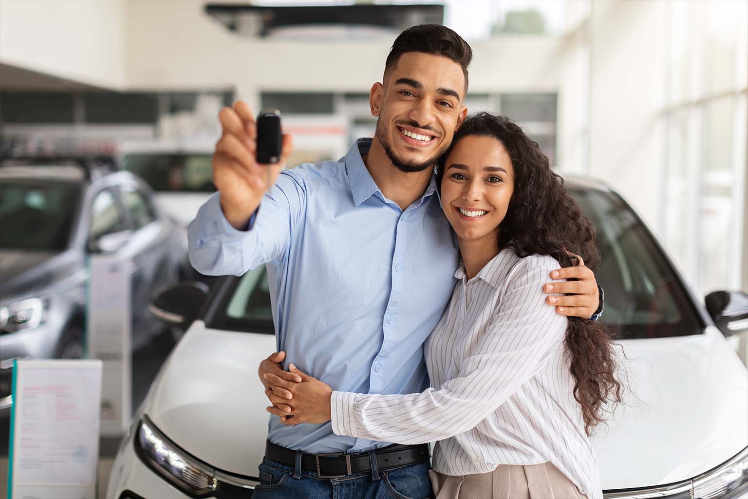 Casal jovem sorridente comemora a conquista de um novo carro branco em uma concessionária, segurando as chaves e ilustrando o benefício do Crédito em Dobro Consórcio Santa Emília.