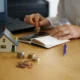 Pessoa calcula despesas ao lado de uma casa em miniatura para avaliar as vantagens do consórcio